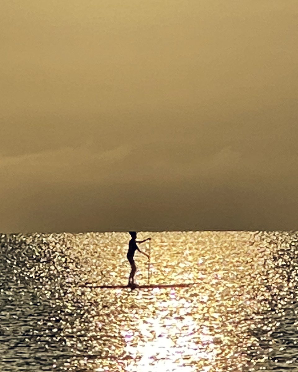A paddleboarder in silhouette, standing steady on calm water at a cloudy sunrise, the sun’s path glittering gold across the sea